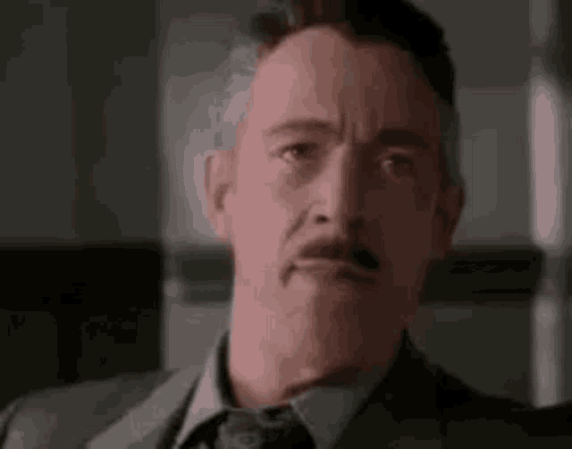 A mustached man stares forward with a serious, uneasy expression.