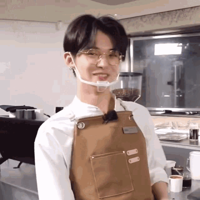 a man wearing glasses and an apron is standing in front of a coffee grinder in a kitchen .