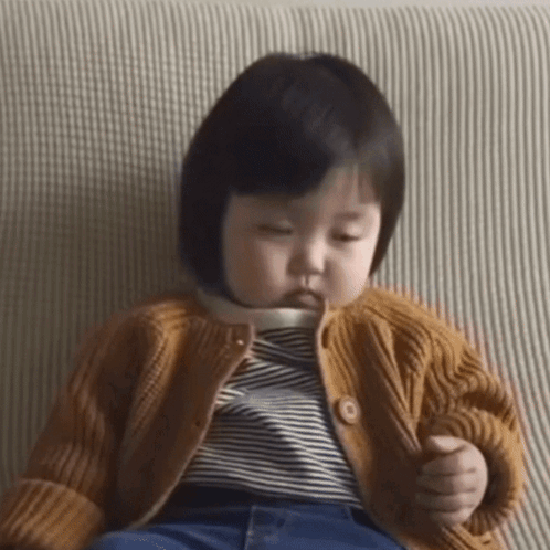 A young child sits sleepily on a sofa holding a small object.