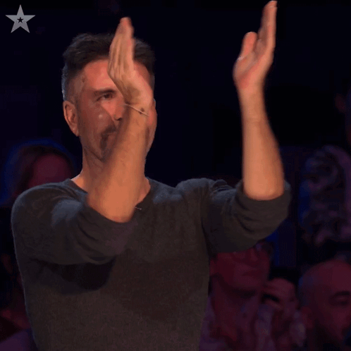 Simon Cowell claps appreciatively in a dark audience setting.