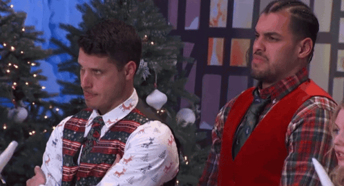 two men standing in front of a christmas tree wearing ugly christmas sweaters