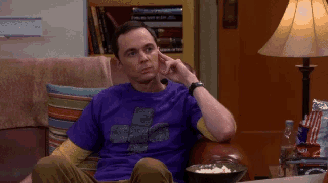 a man in a purple shirt sits on a couch with a bowl of popcorn
