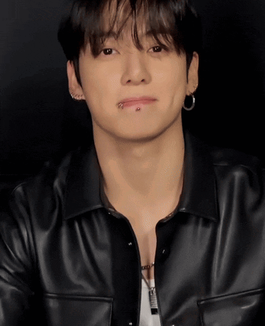 A man in a black leather shirt looks ahead with a restrained smile.