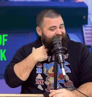 A man at a microphone points to himself with a strained laugh.