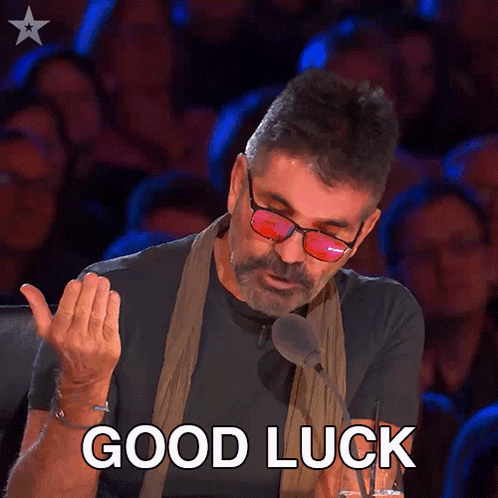 Simon Cowell gestures with a doubtful expression beneath “GOOD LUCK.”