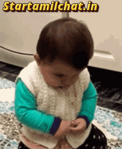 A toddler in a green shirt looks down while standing on a patterned rug.
