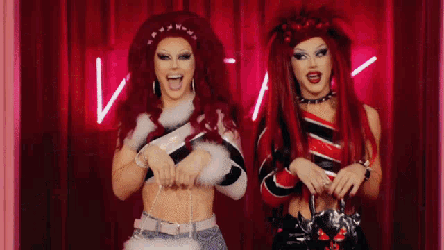 two drag queens are standing next to each other in front of a pink neon sign .
