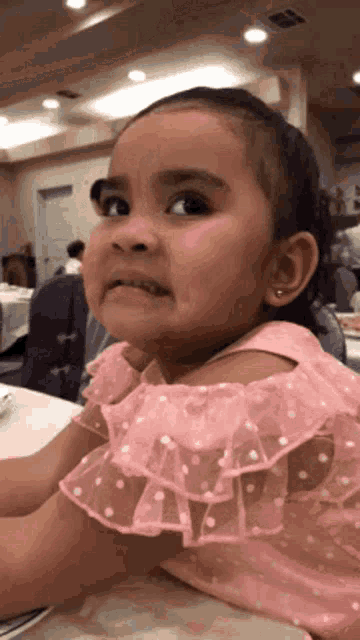 A little girl in a pink ruffled dress looks up warily in a busy room.