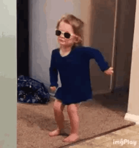A toddler in sunglasses dances indoors.