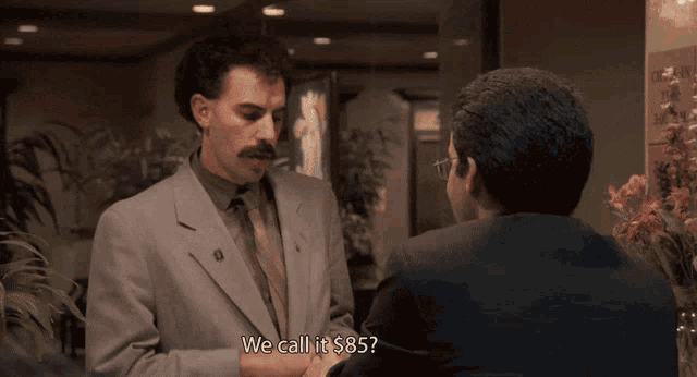 a man in a suit and tie is talking to another man and they both say we call it $ 85