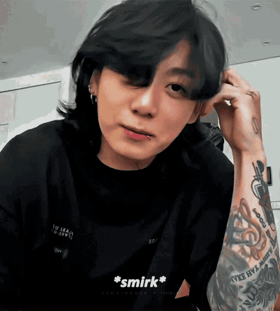 A tattooed man touches his hair and gives a shy smirk.