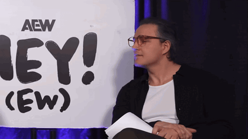 Matt Cardona sits holding papers beside the “HEY! (EW)” sign.