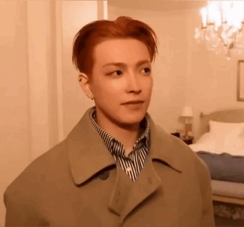 a young man with red hair is standing in a bedroom wearing a coat and striped shirt .
