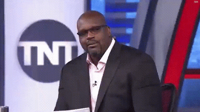 A suited sports commentator looks aside with an amused expression.