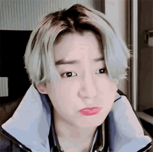 A blond-haired man makes a worried pout while touching the back of his neck.