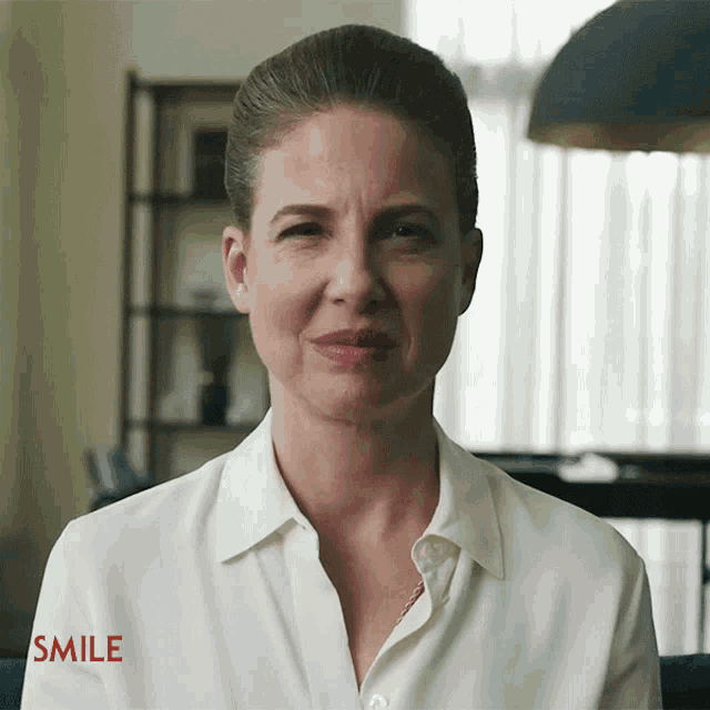 A woman in a white blouse holds an uncomfortable, restrained smile.