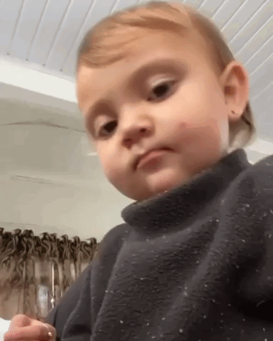 A toddler in a dark sweater gives a small approving thumbs-up look.