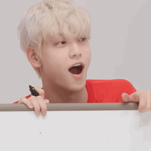 A white-haired performer peers over a sign with a surprised open-mouthed look.
