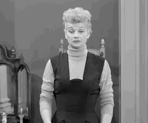 A black-and-white woman stands with a blank expression in a vintage interior.
