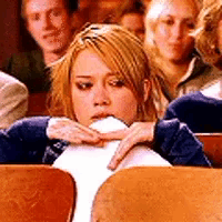 A girl sits at a desk looking uncomfortable while people stand behind her.