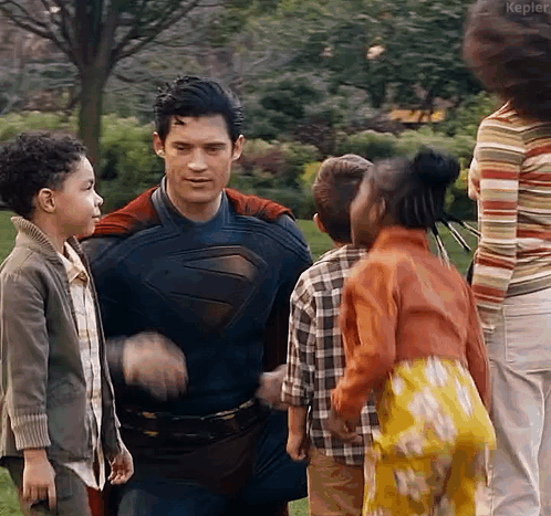 a man in a superman costume is hugging a group of children in a park .
