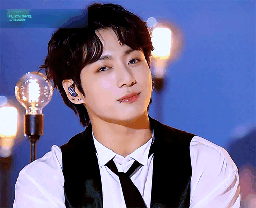 Jungkook smiles softly while wearing a white shirt, black vest, and tie onstage.