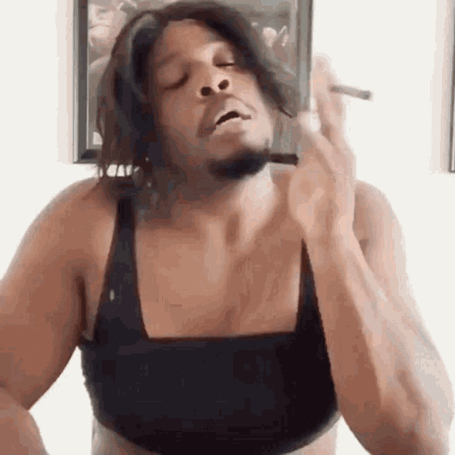 A person in a black tank top takes a drag from a cigarette with a tired expression.