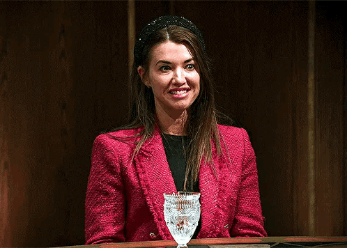 a woman in a red jacket is sitting at a table with a glass of wine and making a funny face .