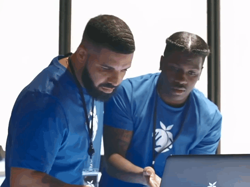 Two men in blue shirts lean over a laptop together.