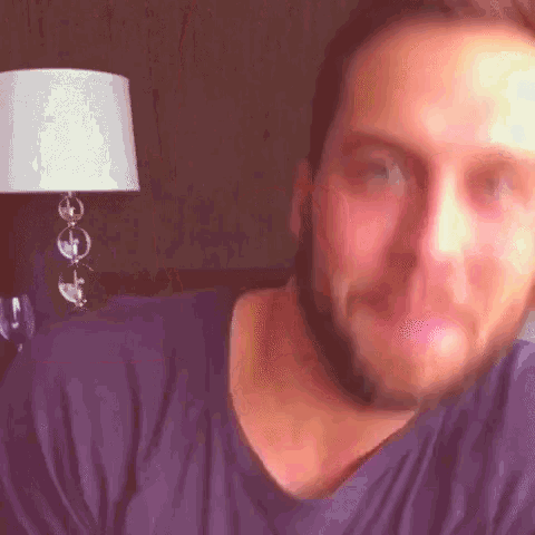 a man with a beard wearing a purple shirt looks at the camera with a lamp in the background