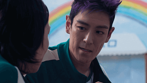 a man with purple hair talks to another man in front of a rainbow