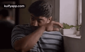 a man in a striped shirt is sitting in front of a window and rubbing his face .