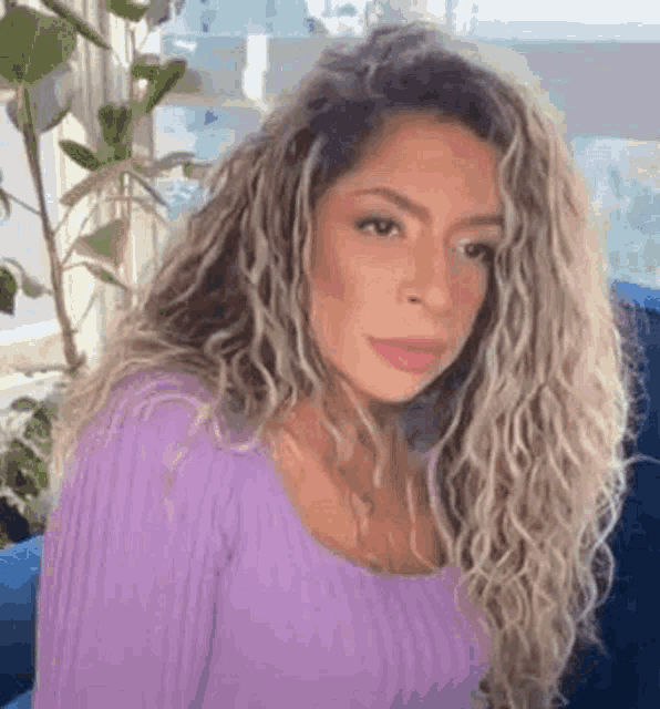 a woman with curly blonde hair is wearing a purple sweater and sitting on a blue couch .