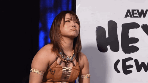 Yuka Sakazaki sits silently in front of a “HEY! (EW)” sign.