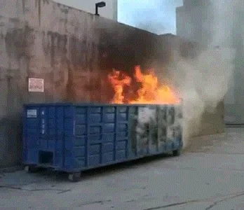 A large dumpster burns intensely beside a building.