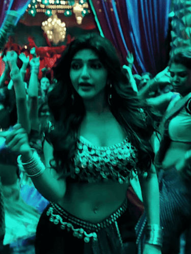 A woman in a jeweled dance costume moves through a teal-lit crowd.