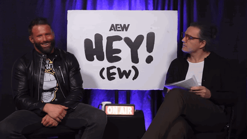 Matt Cardona smiles beside a “HEY! (EW)” sign during an interview.