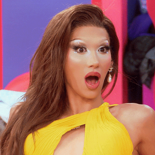 A drag performer in a yellow outfit stares wide-eyed with their mouth open.