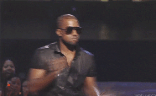 Kanye West in sunglasses makes a quick shrugging gesture onstage.