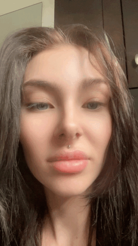 A woman in a vertical selfie video stares forward with a neutral-to-displeased look.