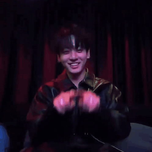 Jungkook smiles and forms a heart gesture with his hands in a dark setting.