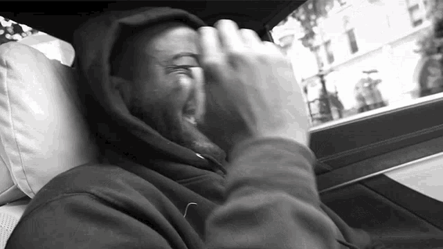 a man in a hoodie is sitting in a car and covering his face with his hand
