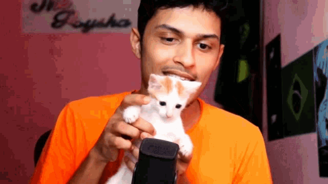 a man in an orange shirt is holding a kitten and a cell phone