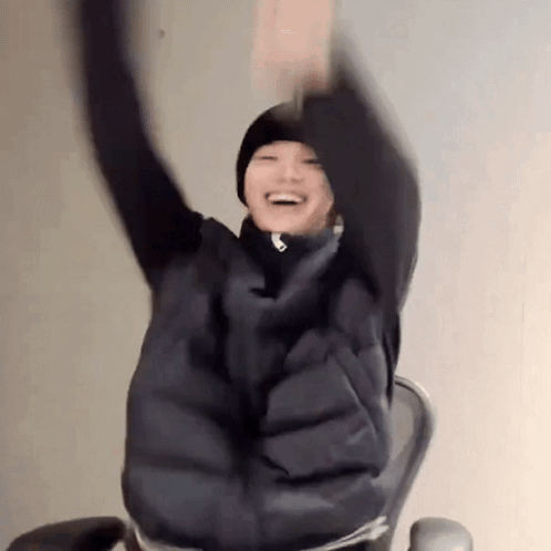 A person in a dark puffer coat raises both arms in excitement while seated.