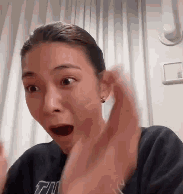 A woman gasps and lifts a hand toward her cheek in surprise.