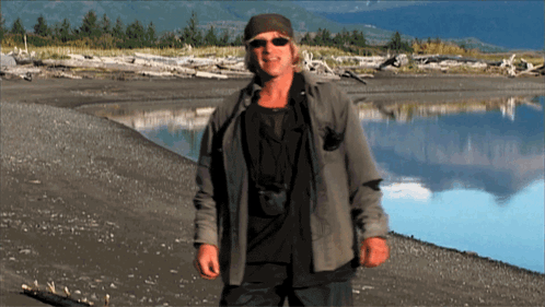 a man wearing sunglasses and a hat stands on a beach next to a body of water