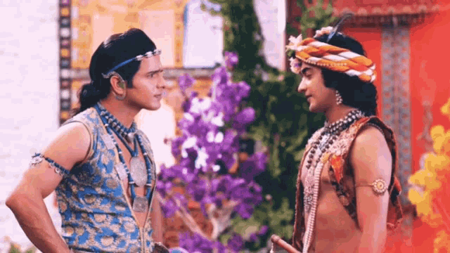 two men are standing next to each other and talking to each other in front of flowers .