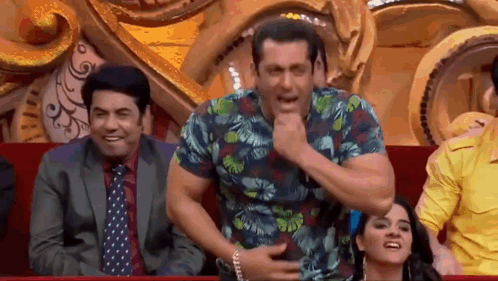 a man in a hawaiian shirt is sitting in a theatre with other people and making a funny face .