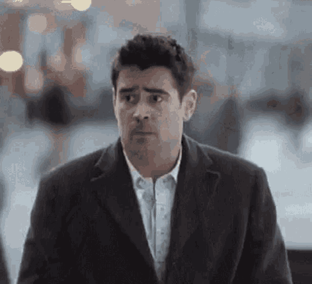 A man in a dark suit looks uncertain in a softly lit public space.