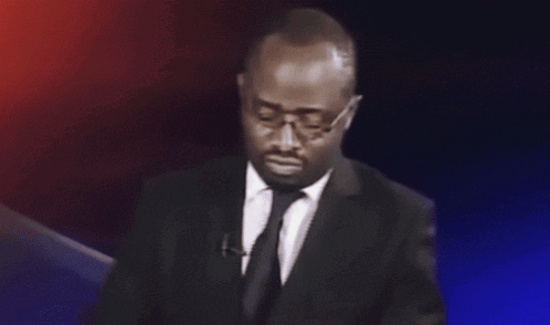 A suited man at a podium looks aside in a questioning reaction.